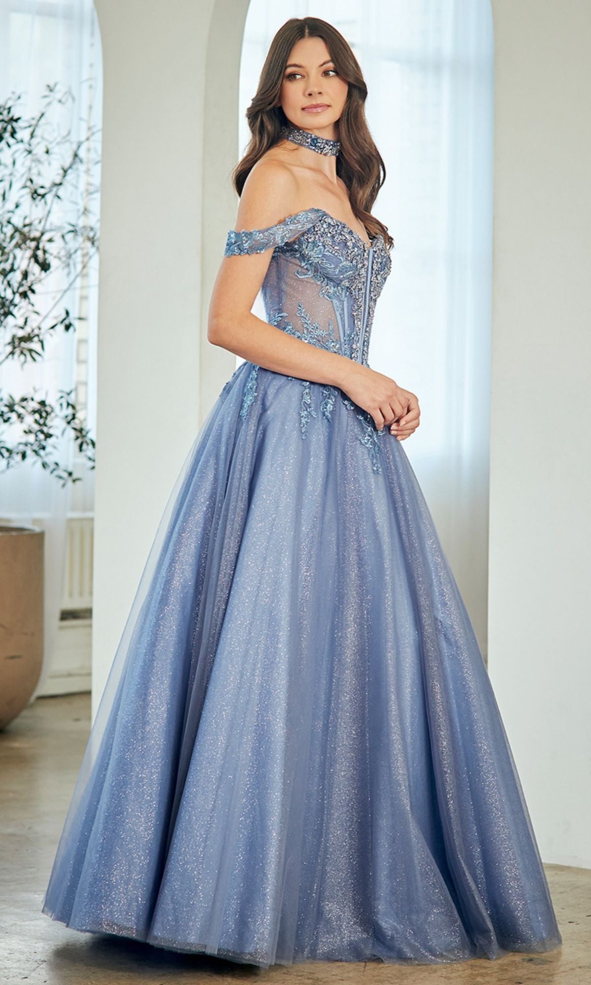 A woman in the Amelia Couture 8065 long prom dress, featuring a shimmery off-the-shoulder design with embroidered details and a structured corset bodice, stands indoors near a window.